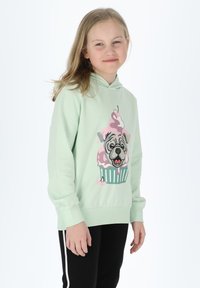 Felpa verde menta con un cane dei cartoni animati che presenta un design di cupcake e gelato. Tessuto morbido, vestibilità rilassata e polsini a coste. Pantaloni neri.