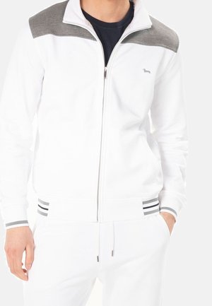 Homme portant une veste blanche à fermeture éclair avec des panneaux gris aux épaules et des poignets rayés, associée à un pantalon blanc, mains dans les poches.