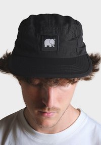 Čierna čiapka s okrúhlou korunou, ktorá má vpredu štvorcový nášivku s bielym logom slona. Vizír je plochý s detailným prešívaním.