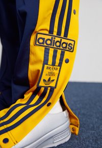Adidas Trainingshose in Gelb und Marineblau, mit drei charakteristischen Streifen, Marken-Logo-Patches, Druckknöpfen und einer lässigen Passform.