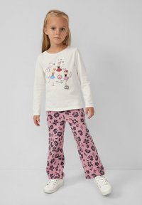 Ragazzina in una camicia bianca a maniche lunghe con stampa di artisti circensi, pantaloni floreali rosa e sneakers bianche, in piedi contro uno sfondo neutro.