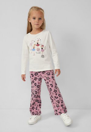 Meisje in een wit shirt met lange mouwen en een circusartiestenprint, roze bloemenbroek en witte sneakers, staand tegen een effen achtergrond.