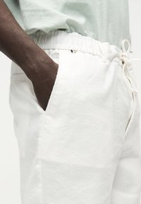 Άτομο που φοράει άσπρο παντελόνι με κορδόνι και ελαστική μέση, με σκουρόχρωμο χέρι στερεωμένο στην εμπρόσθια τσέπη, φορώντας ανοιχτό πράσινο πουκάμισο.