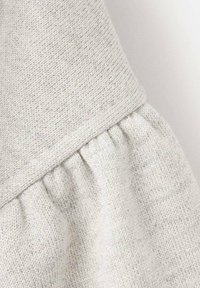 Světlá šedá pletená látka s texturovaným povrchem. Má nashromážděný šev dole a hladký, zalomený horní okraj.