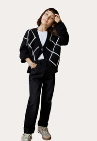 Schwarze Strickjacke mit weißem Diamantmuster, Knöpf front, getragen über einem weißen Hemd, kombiniert mit schwarzen Hosen und beigen Sneakers.