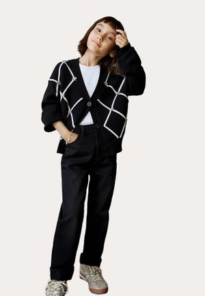 Zwarte cardigan met wit ruitpatroon, knopen aan de voorkant, gedragen over een wit shirt, gecombineerd met zwarte broek en beige sneakers.