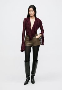 Vínový krajkový top s volánky vpředu a zvonovými rukávy; v kombinaci s olivově zelenými koženými šortkami a černými kozačkami na podpatku po kolena.