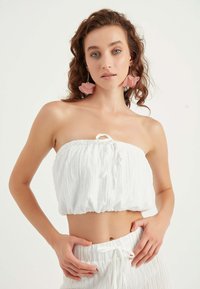 Top corto bianco con tessuto testurizzato e una cravatta regolabile, abbinato a pantaloni bianchi con vita elasticizzata. La modella indossa orecchini floreali rosa.