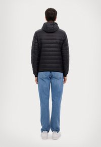 Giacca nera imbottita con cappuccio, caratterizzata da un design trapuntato e polsini elasticizzati, abbinata a jeans in denim blu e sneakers bianche.