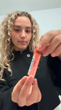 Produkt do ust w kolorze brzoskwiniowym trzymany w ręku, o cylindrycznym kształcie z ciemniejszym dnem i etykietą z boku. Tekstura wydaje się gładka.