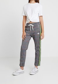 Grijze geruite broek met rode en zwarte accenten, voorzien van een neon groene zijstreep en een taille met trekkoord, gecombineerd met een witte geknoopte T-shirt.