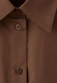 Chemise marron à boutons avec col pointu et trois boutons ronds visibles sur un tissu lisse en gros plan.