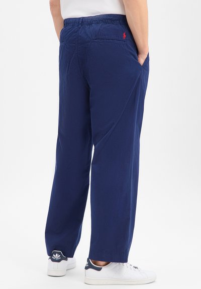 Personne portant un pantalon décontracté bleu marine avec une taille élastique et des baskets blanches, debout les mains dans les poches, dos tourné.