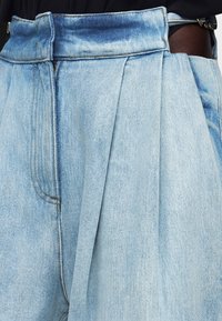 Ανοιχτό μπλε πλισέ τζιν παντελόνι που φοριέται από άτομο με μαύρο τοπ, εμφανίζοντας λεπτομέρειες στη μέση, το φερμουάρ και την πλαϊνή τσέπη.