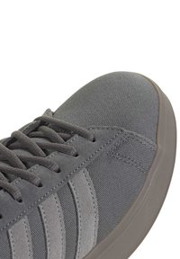 Ingrandimento dell'area della punta di una sneaker grigia con texture in tessuto, strisce in suede, lacci coordinati e suola in gomma beige su sfondo bianco.