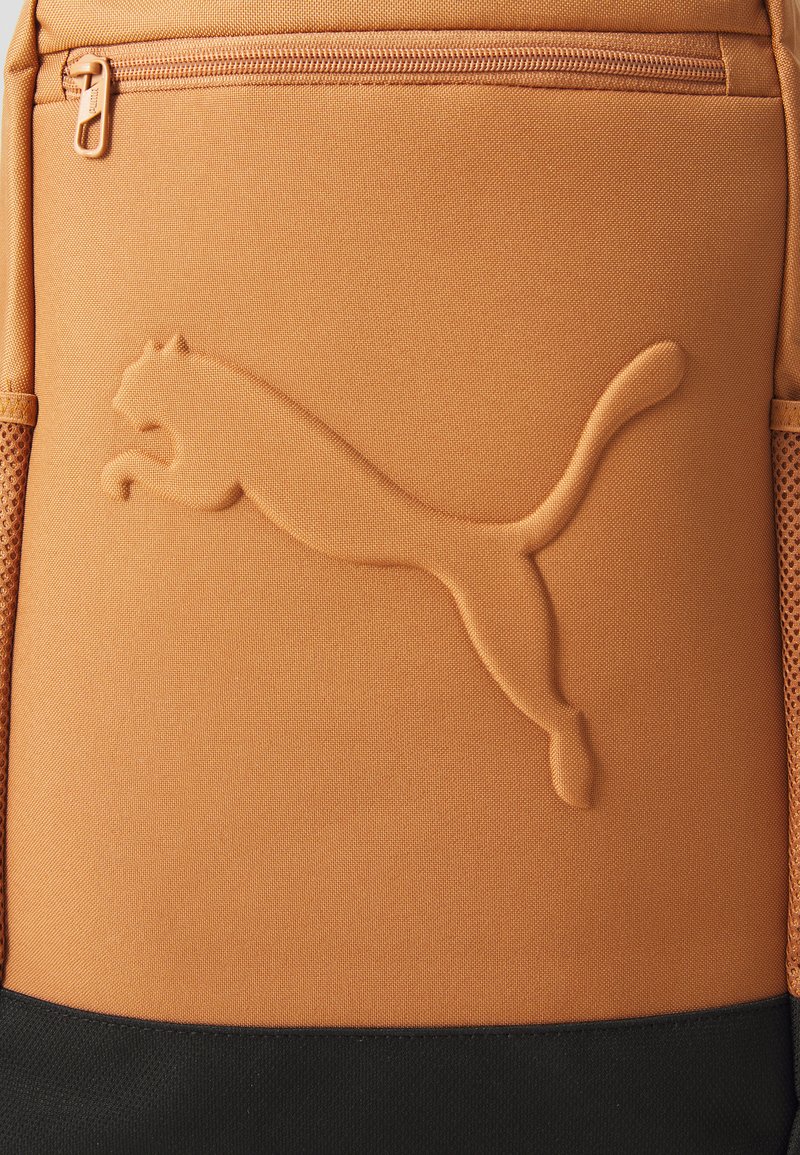 Mochila de tela naranja con superficie texturizada, que presenta un logotipo en relieve destacado y un panel inferior negro. Incluye un bolsillo con cremallera.