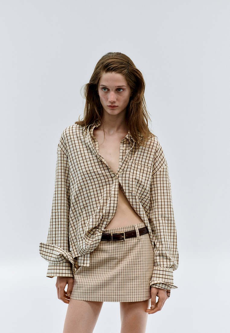 Jeune femme portant une chemise à carreaux ample sur une mini-jupe assortie à carreaux avec une ceinture foncée, debout devant un fond clair uni.