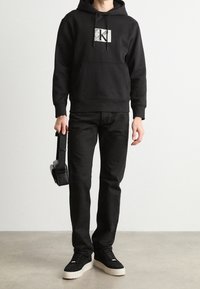 Sudadera con capucha negra con un logotipo gráfico, combinada con jeans negros y zapatillas negras con suela blanca. El modelo sostiene unos auriculares negros.