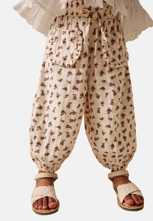 Kind trägt beige Hose mit kleinem rosa Blumenmuster und Rüschentaschen, kombiniert mit cremefarbenen gewebten Sandalen.