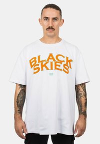 Vit t-shirt i bomull med fet orange och grön text "BLACK SKIES". Korta ärmar, lös passform, rak nederkant. Anmärkningsvärda tatueringar på synliga armar.