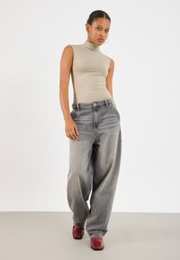 Haut sans manches beige à col montant associé à un jean gris délavé oversized et des chaussures à talons carrés rouges brillantes. Design simple, coupe décontractée.