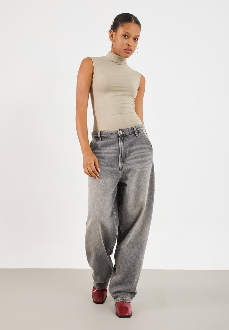Haut sans manches beige à col montant associé à un jean gris délavé oversized et des chaussures à talons carrés rouges brillantes. Design simple, coupe décontractée.