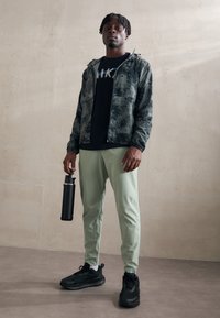 Sort og grøn mønstrede zip-up jakke over en sort T-shirt, parret med lyse grønne bukser og sorte sportsko. Holder en sort vandflaske.