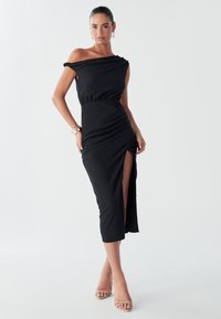 A woman poses in a fitted black dress with a high slit, off-the-shoulder design, and strappy heels, set against a plain background.