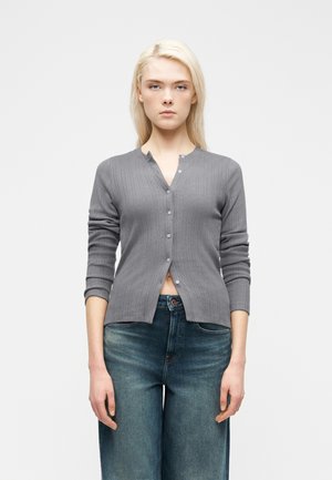 Femme aux cheveux blonds portant un cardigan gris boutonné et un jean bleu, debout devant un fond clair uni.