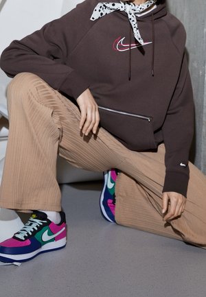 Osoba ubrana w brązową bluzę z kapturem z podwójnym logo Nike, beżowe prążkowane spodnie, chustę w groszki na szyi oraz kolorowe buty Nike, przykuca w pomieszczeniu.