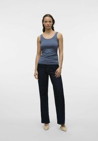 Débardeur sans manches bleu, design ajusté, associé à un jean droit foncé. Le mannequin porte des chaussures à talons pointus beiges, se tenant devant un fond blanc.
