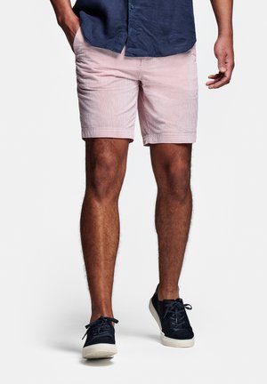 Man die een marineblauw shirt, lichtroze gestreepte korte broek en zwarte sneakers draagt, staand met één hand in de zak en de andere langs het lichaam.