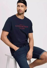 Granatowa koszulka z okrągłym dekoltem z czerwonym nadrukiem "CROSS JEANS LE DENIM POUR LA VIE", w zestawieniu z ciemnymi szortami dżinsowymi. Gładka tekstura bawełny.