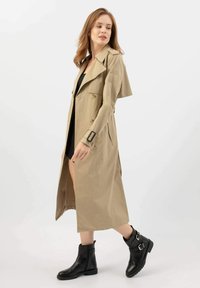 Beżowy płaszcz w trenczowym stylu z paskiem w talii, podwójnym zapięciem i pagonami na ramionach, w połączeniu z czarnymi botkami za kostkę z klamrami.