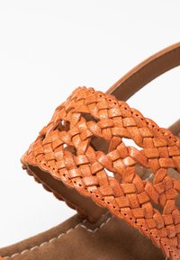 Braided orange leather strap with cut-out pattern, stitched brown suede base, curved design. Focus on texture and color contrasts.