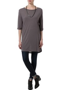 Grå tunika med trekvartsärmar, mjuk textur, asymmetrisk fåll, sidoficka; kombinerad med mörka jeans och svarta ankelstövlar.