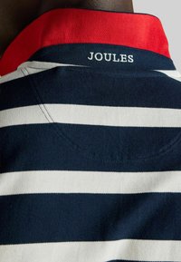 Jūras un baltas svītrains polo krekls ar sarkanu apkakli. Zīmola nosaukums "JOULES" ir izšūts uz apkakles. Mīksts kokvilnas materiāls.