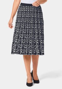 Jupe longueur genou, bleu marine avec des motifs fléchés blancs ; design plissé, taille élastique, en tissu léger, portée avec des talons noirs.