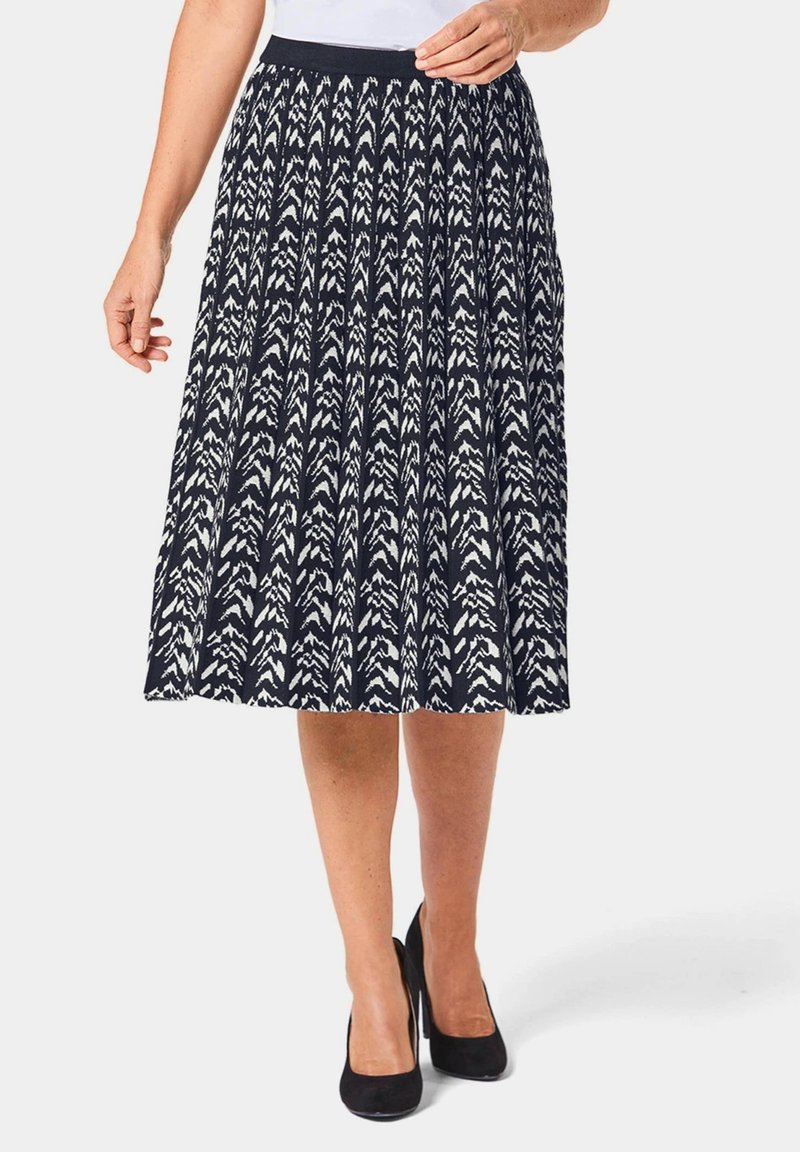 Jupe longueur genou, bleu marine avec des motifs fléchés blancs ; design plissé, taille élastique, en tissu léger, portée avec des talons noirs.