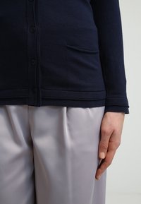 Cardigan blu navy con bottoni, dotato di tasca e orlo a coste. Abbinato a pantaloni in satin grigio chiaro, creando un contrasto di texture morbida.