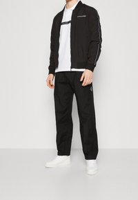 Černá bomber bunda se zipem, bílý detail loga, v kombinaci s bílým polo tričkem, černými kalhotami a bílými teniskami. Hladká textura, ležérní styl.