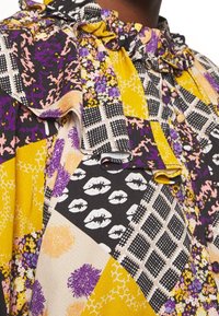 Blouse multicolore avec un col à volants, présentant des motifs de lèvres, floraux et géométriques dans des tons de violet, jaune, noir et rose.