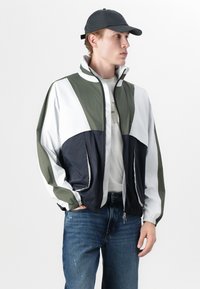 Fargeblokkerte vindjakker i olivengrønn, marineblå og hvit med elastiske mansjetter, hel glidelås og sidelommer. Brukt med blå jeans.