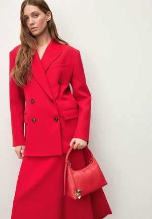 Punane topeltkinnisega bleiser kuldsete nööpidega, mida kannab sobiva pikkade seeliku komplekt. Hoides käes väikest punast nahast käekotti kuldsete detailidega.