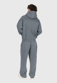 Person mit lockigem, dunklem Haar, der einen grauen Kapuzenpullover und passende Hosen trägt, mit den Händen in den Taschen steht und von hinten schaut.