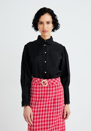 Blusa negra con cuello volante y tapeta delantera con botones blancos; combinada con una falda a cuadros rojos y hebilla decorativa en el cinturón.