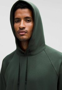 Vihreä huppari, jossa on nyörit ja suuri huppu. Sileä rakenne ja raglanhihat luovat saumattoman muotoilun. Mallin etunäkymä.