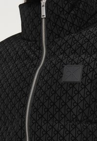 Czarna kurtka puchowa z teksturowanym wzorem, wysoki kołnierz i srebrny zamek błyskawiczny. Ma kwadratową naszywkę z logo oraz pikowany wzór.