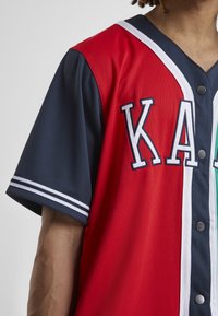 Piros és tengerészkék baseball mez fehér díszítéssel, gombos elülső rész, és a "KAY" felirat szépen kiemelve a mellkason.