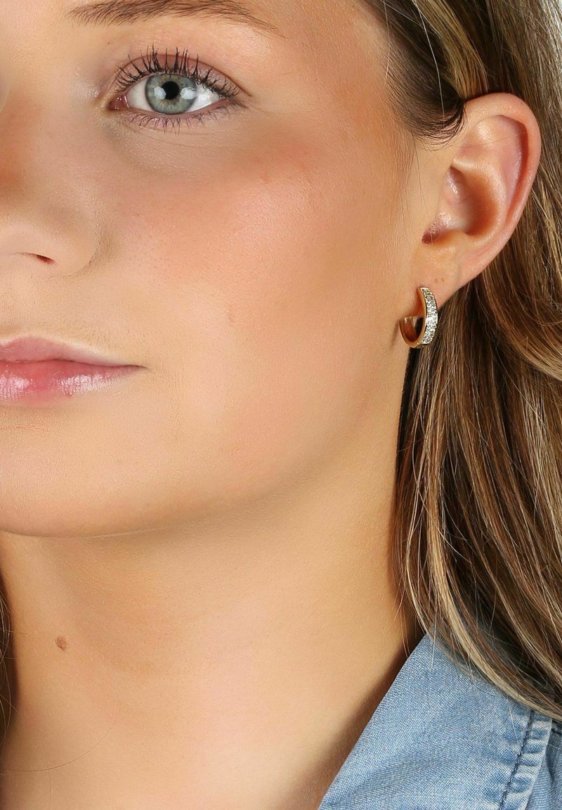 Guldtonad hoopsnäcka med en böjd design, dekorerad med klara rhinestones längs den främre kanten, mot en ljus hudton.